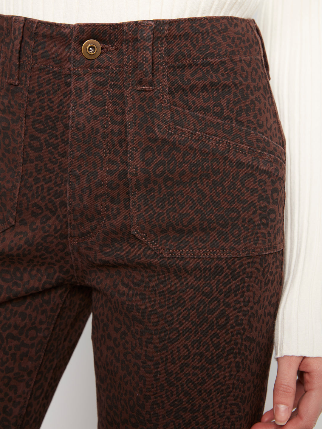Close-up of dark brown leopard-print jeans worn by a model, showing the back pocket, button, and part of the white ribbed top and hand.