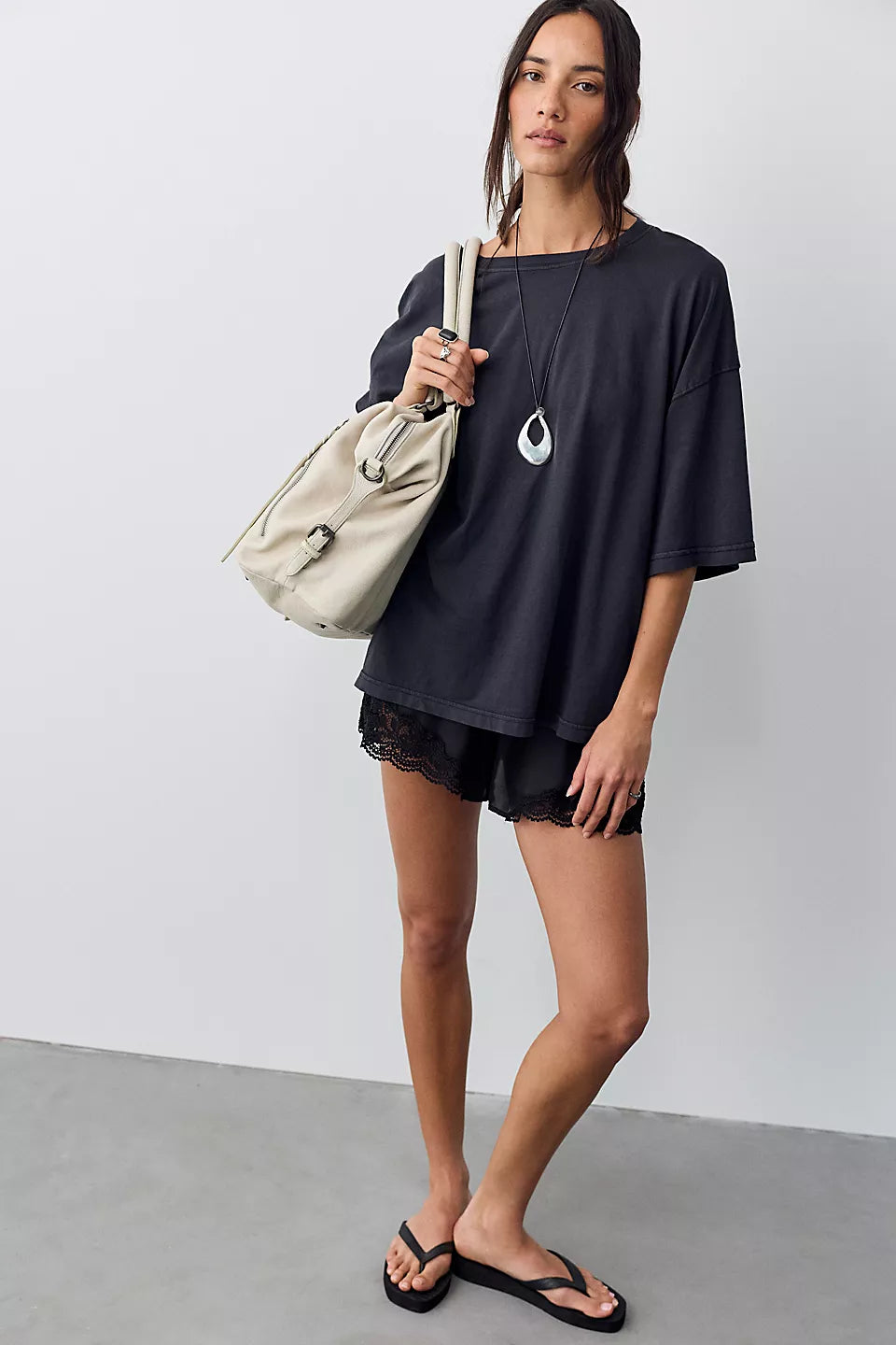 A woman modeling an oversized black t-shirt with a white graphic print, black shorts, black flip-flops, and carrying a beige shoulder bag against a plain studio background.