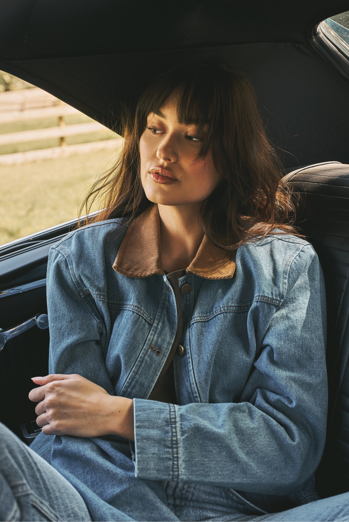 A woman with dark hair sits in the front passenger seat of a car, wearing a light blue denim jacket with a brown corduroy collar.