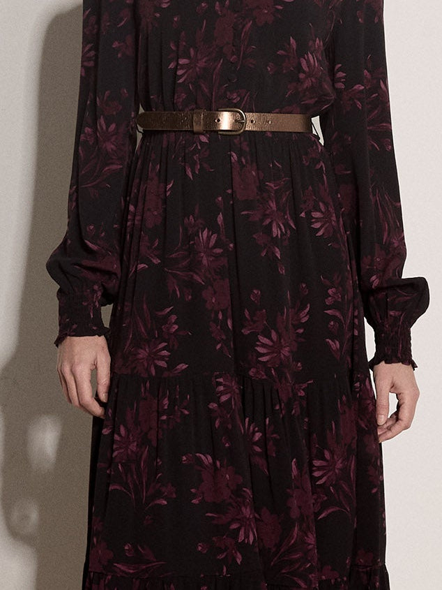 A model stands against a plain off-white wall wearing a long-sleeved midi dress with a dark floral pattern, a thin metallic gold waist belt, and dark brown ankle boots.