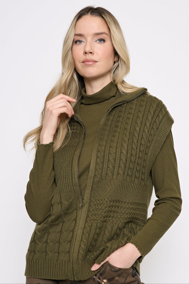 A woman modeling an olive green cable-knit zip-front vest over a matching olive green mock-neck top, paired with brown patterned pants, against a plain light gray studio background.