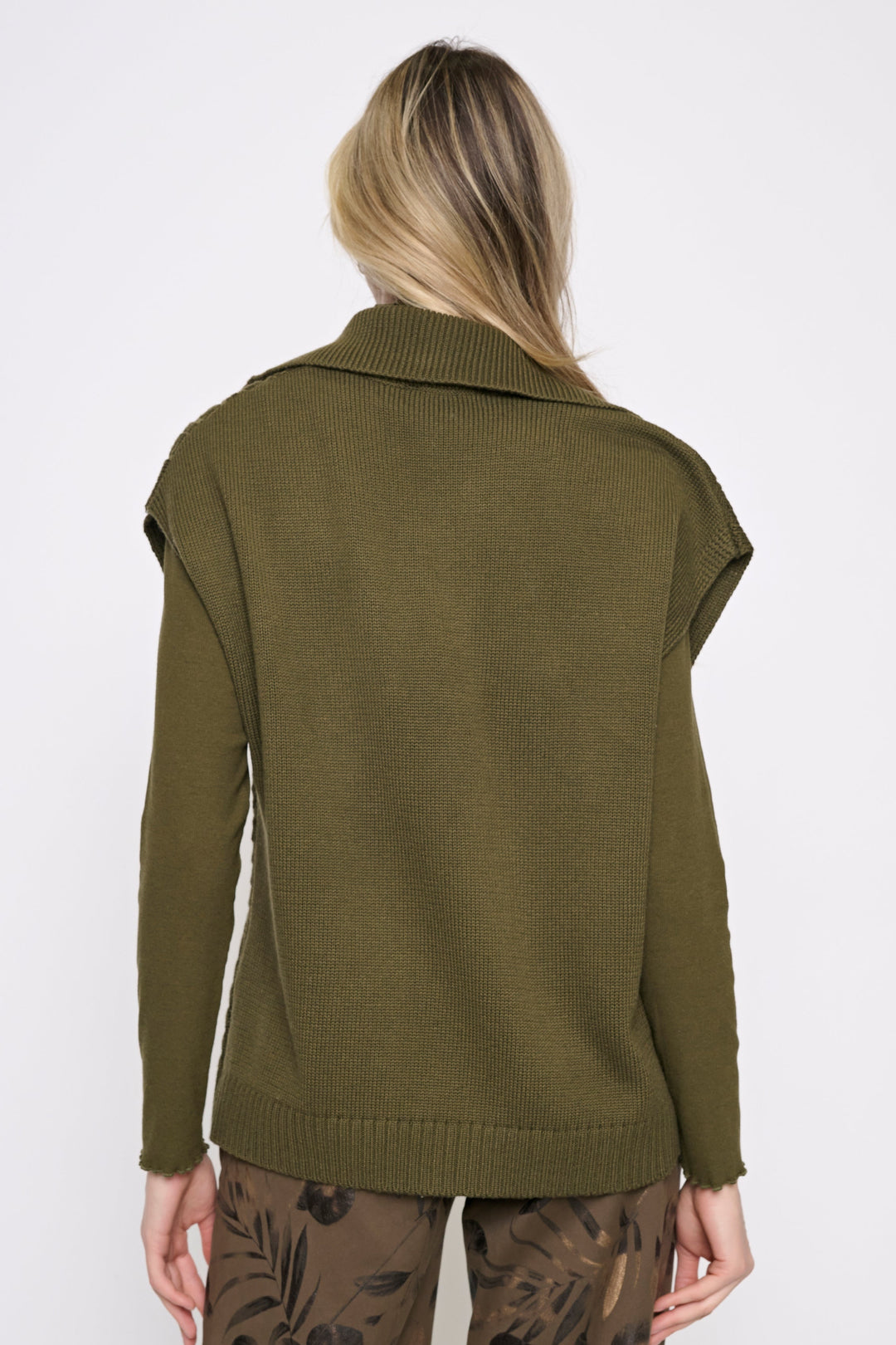 A woman viewed from the back wearing an olive green knit sweater with a wide collar and cropped sleeves, paired with brown patterned pants, against a plain white studio background.