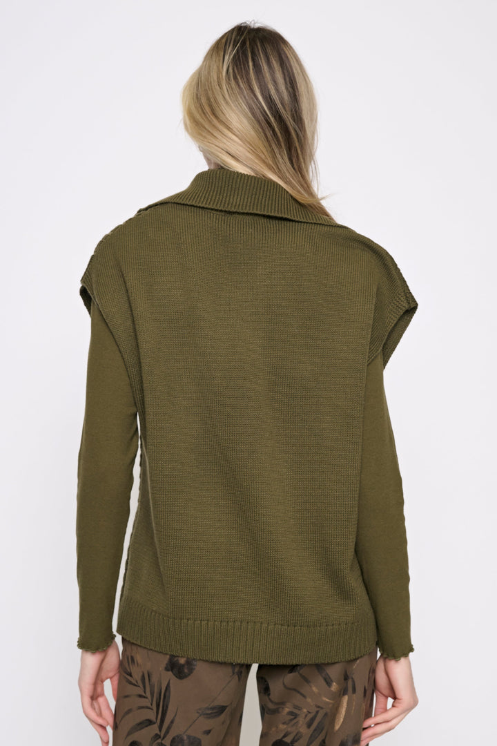 A woman viewed from the back wearing an olive green knit sweater with a wide collar and cropped sleeves, paired with brown patterned pants, against a plain white studio background.