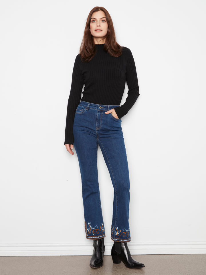 A woman stands against a white wall wearing a black ribbed mock-neck long-sleeve top, blue high-waisted flare jeans with floral embroidery at the hem, and black ankle boots.