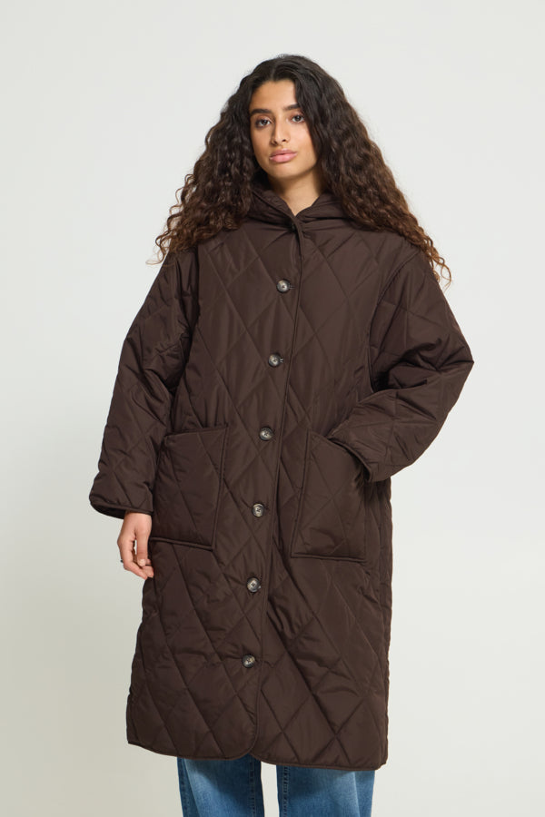A woman with long curly dark hair wearing a dark brown diamond-quilted hooded coat with front buttons, paired with blue jeans, standing against a light gray studio background.