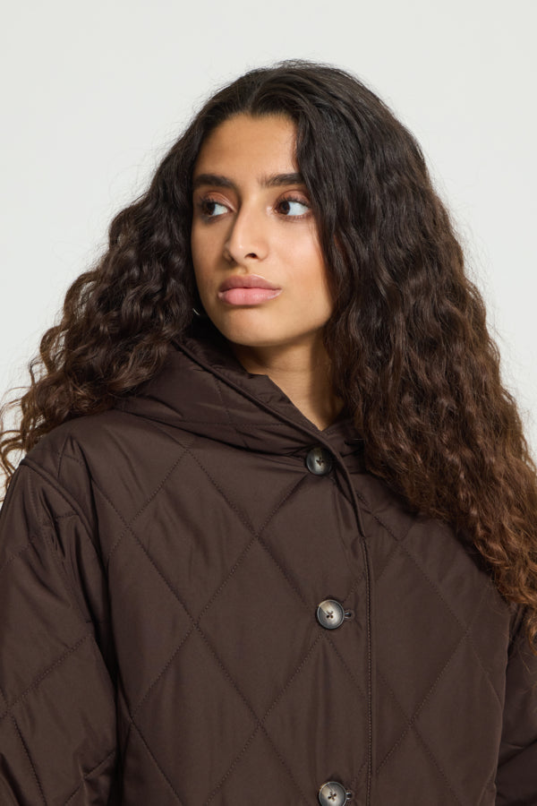 A woman with long, curly dark brown hair wearing a dark brown diamond-quilted jacket with a high collar and silver buttons, photographed against a plain light gray background.