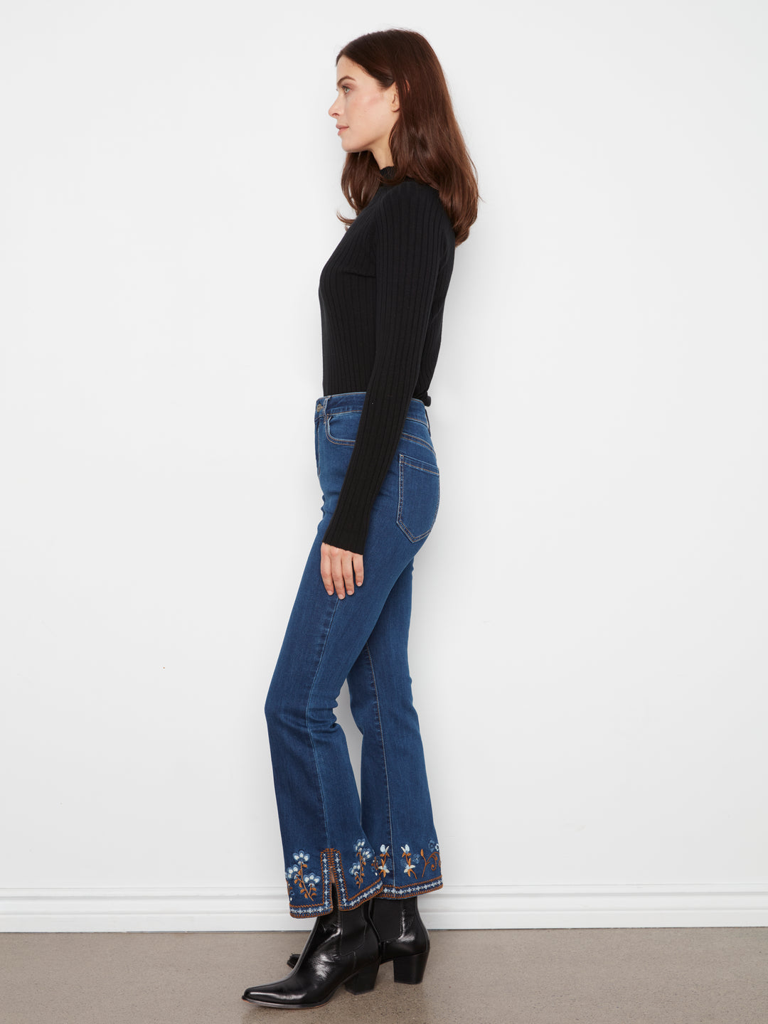 A woman shown in profile wearing a black ribbed long-sleeve top, blue flared jeans with floral embroidery near the hems, and black ankle boots, standing against a plain white wall.
