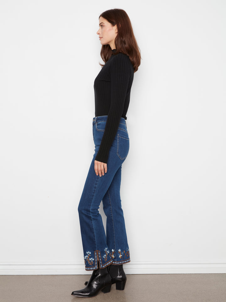 A woman shown in profile wearing a black ribbed long-sleeve top, blue flared jeans with floral embroidery near the hems, and black ankle boots, standing against a plain white wall.