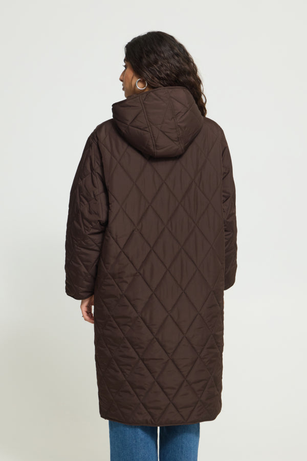 A woman viewed from the back wearing a long quilted brown hooded coat over blue jeans, photographed against a plain light gray studio background.