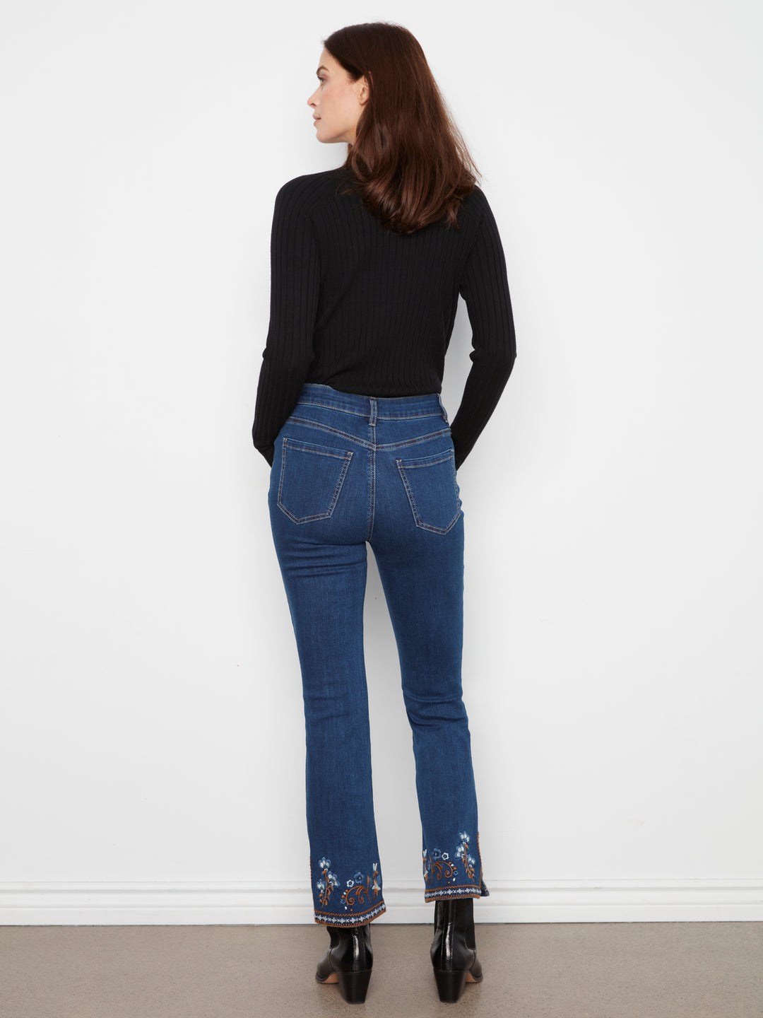 A woman is shown from behind wearing blue flare jeans with floral embroidery at the hem, a black long-sleeve top, and black boots, standing against a white wall.