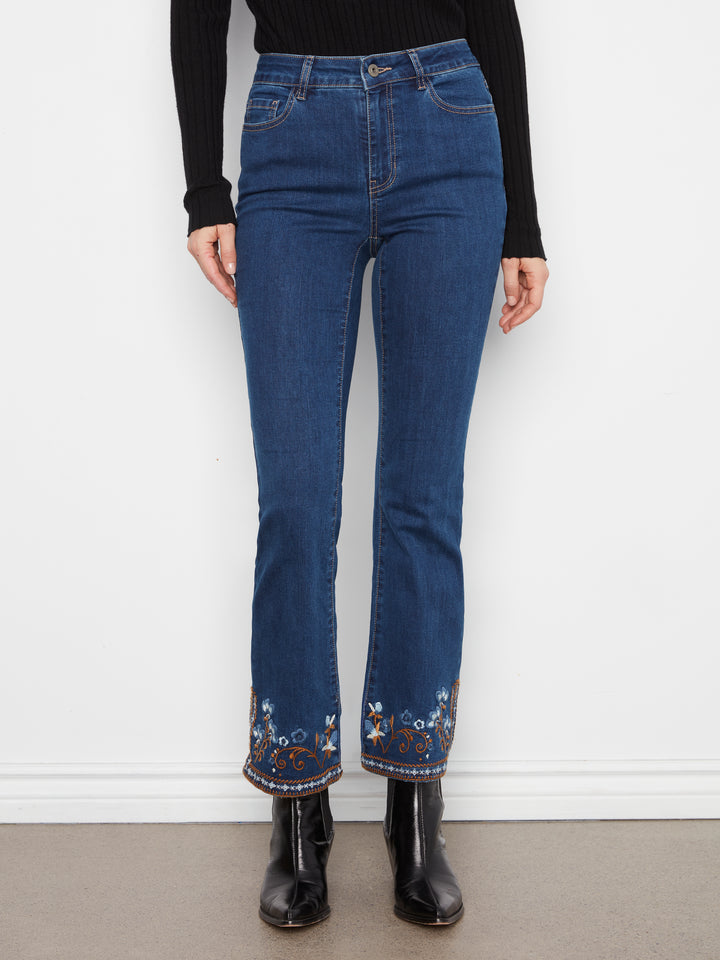 A person wearing blue denim jeans with floral embroidery near the hem, paired with a black long-sleeve top and black ankle boots, standing against a white wall.