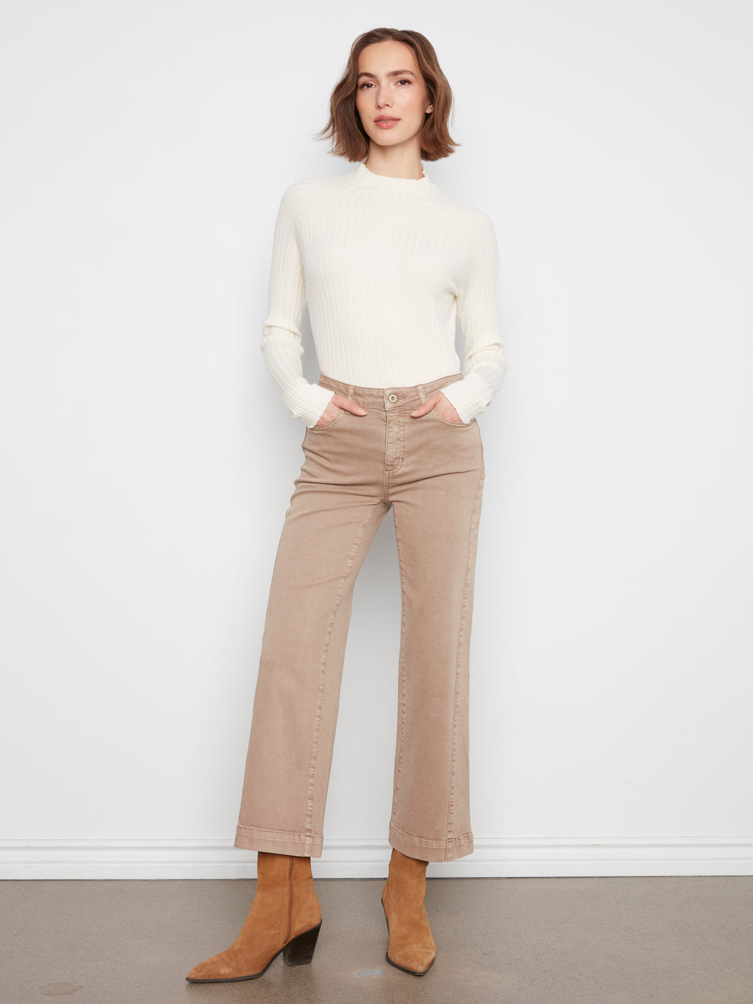 A woman modeling a cream mock-neck knit top and beige wide-leg cropped jeans with tan ankle boots, standing against a white wall.