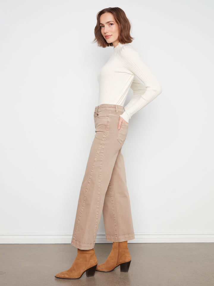 A woman stands in profile wearing a fitted white mock-neck long-sleeve top, beige wide-leg cropped jeans with decorative stitching, and tan ankle boots against a plain white wall and gray floor.