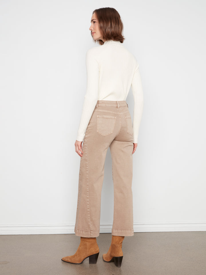 A woman is shown from the back wearing beige wide-leg cropped jeans, a cream mock-neck long-sleeve top, and brown suede ankle boots against a white wall and gray floor.