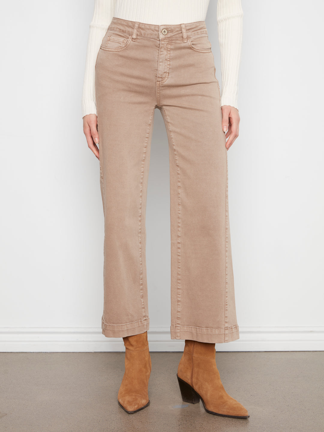 A model is shown from the waist down wearing beige wide-leg cropped jeans, a white ribbed long-sleeve top, and tan ankle boots, standing against a white wall.