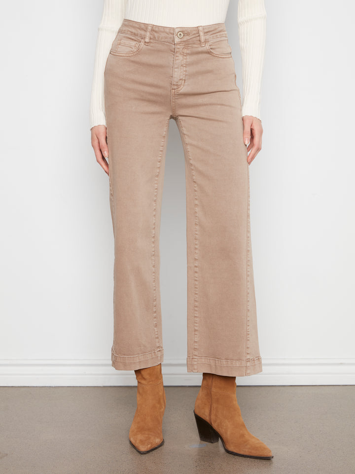 A model is shown from the waist down wearing beige wide-leg cropped jeans, a white ribbed long-sleeve top, and tan ankle boots, standing against a white wall.