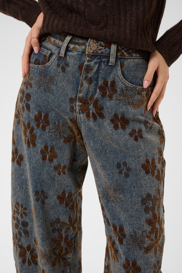 Close-up of a person wearing blue denim jeans with an allover brown floral pattern, paired with a dark brown ribbed sweater against a light gray studio background.