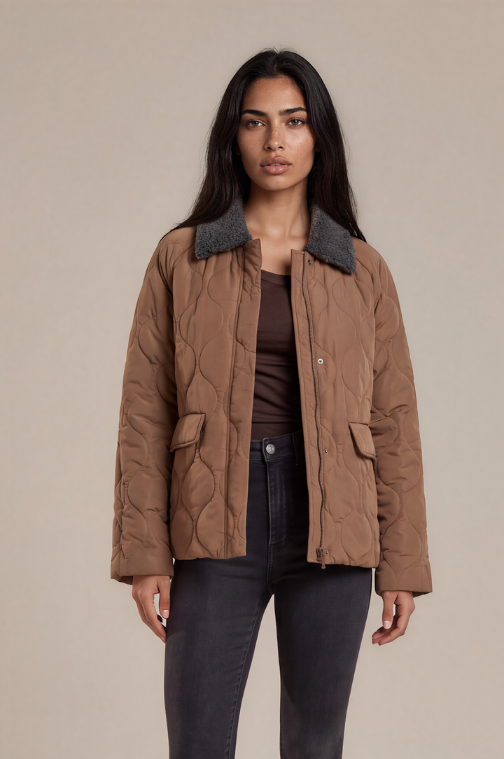 A woman modeling a brown quilted jacket with a dark gray collar, paired with a brown top, dark gray skinny jeans, and black ankle boots, against a solid beige studio background.