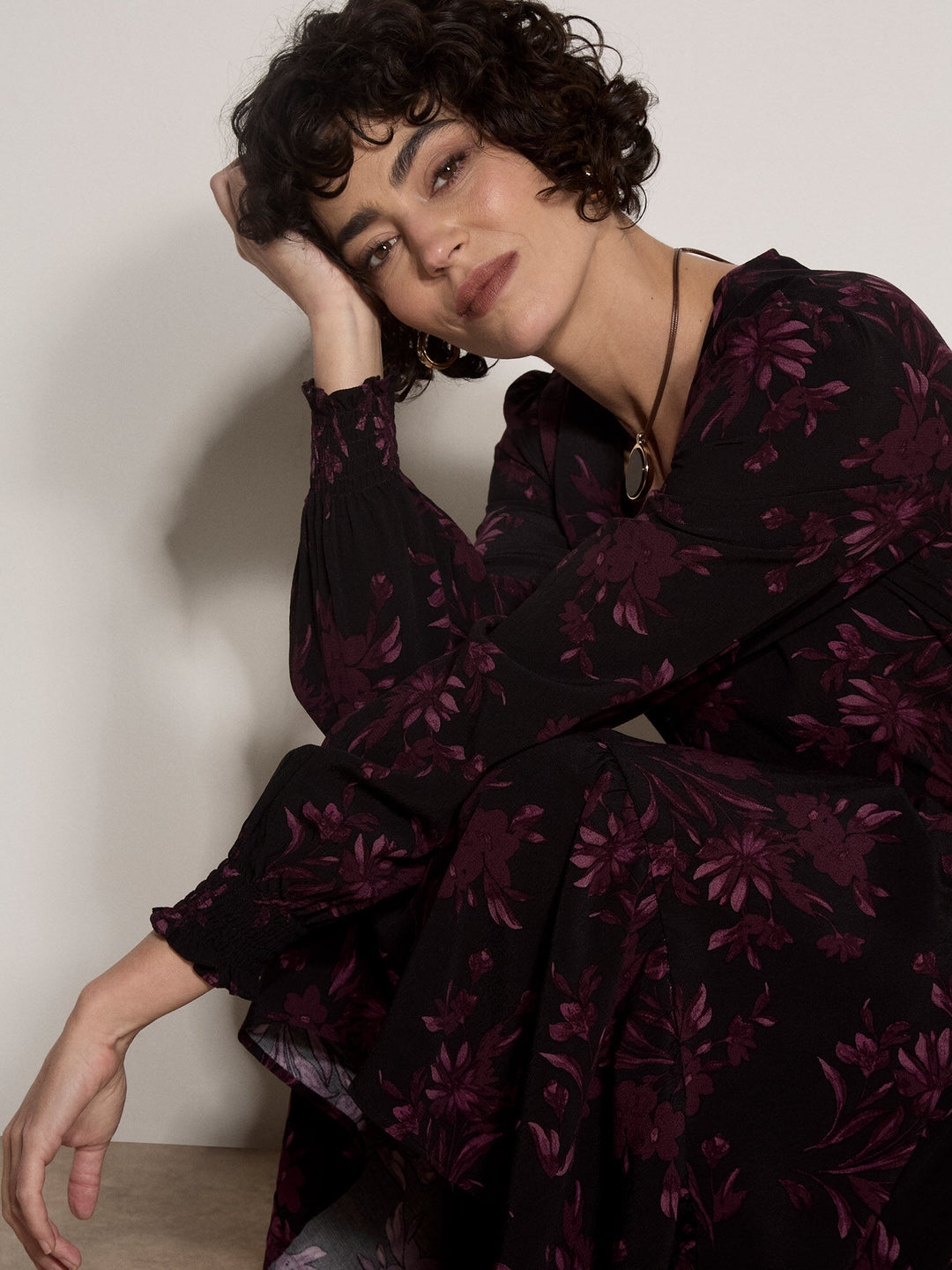 A woman with short curly dark hair wearing a black long-sleeve blouse with a dark purple floral pattern, posing against a plain off-white wall.