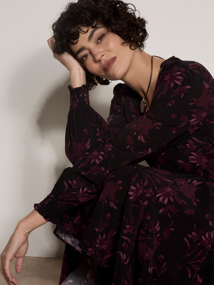 A woman with short curly dark hair wearing a black long-sleeve blouse with a dark purple floral pattern, posing against a plain off-white wall.