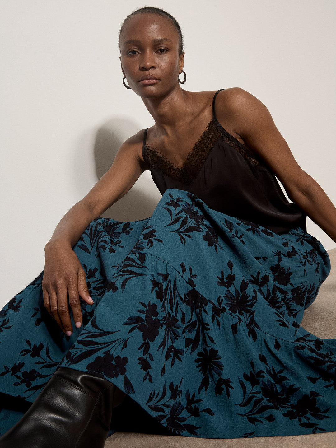 A model wearing a black camisole top and a teal blue maxi skirt with a black floral print, sitting against a plain off-white wall.