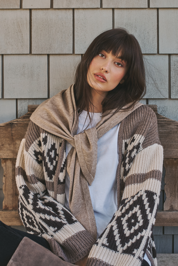 A woman with dark hair and bangs sits on a wooden bench wearing a beige knit cardigan with a brown and cream geometric pattern, a light brown knit wrap, and a white t-shirt against a gray shingled wall.