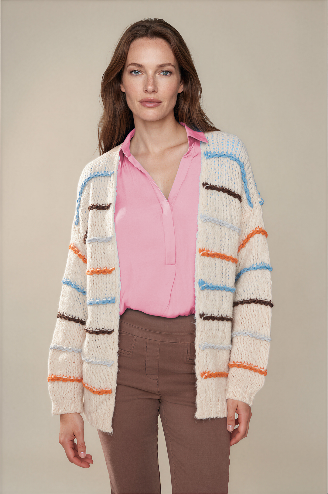 A woman modeling a chunky knit cardigan with horizontal stripes in light blue, orange, brown, and dark brown over a pink blouse and brown pants, set against a solid beige studio background.