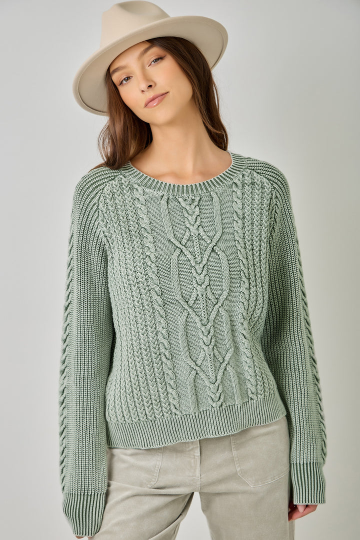 A woman wearing a sage green cable-knit sweater, light beige pants, and a beige wide-brim hat, photographed against a neutral light gray studio background.
