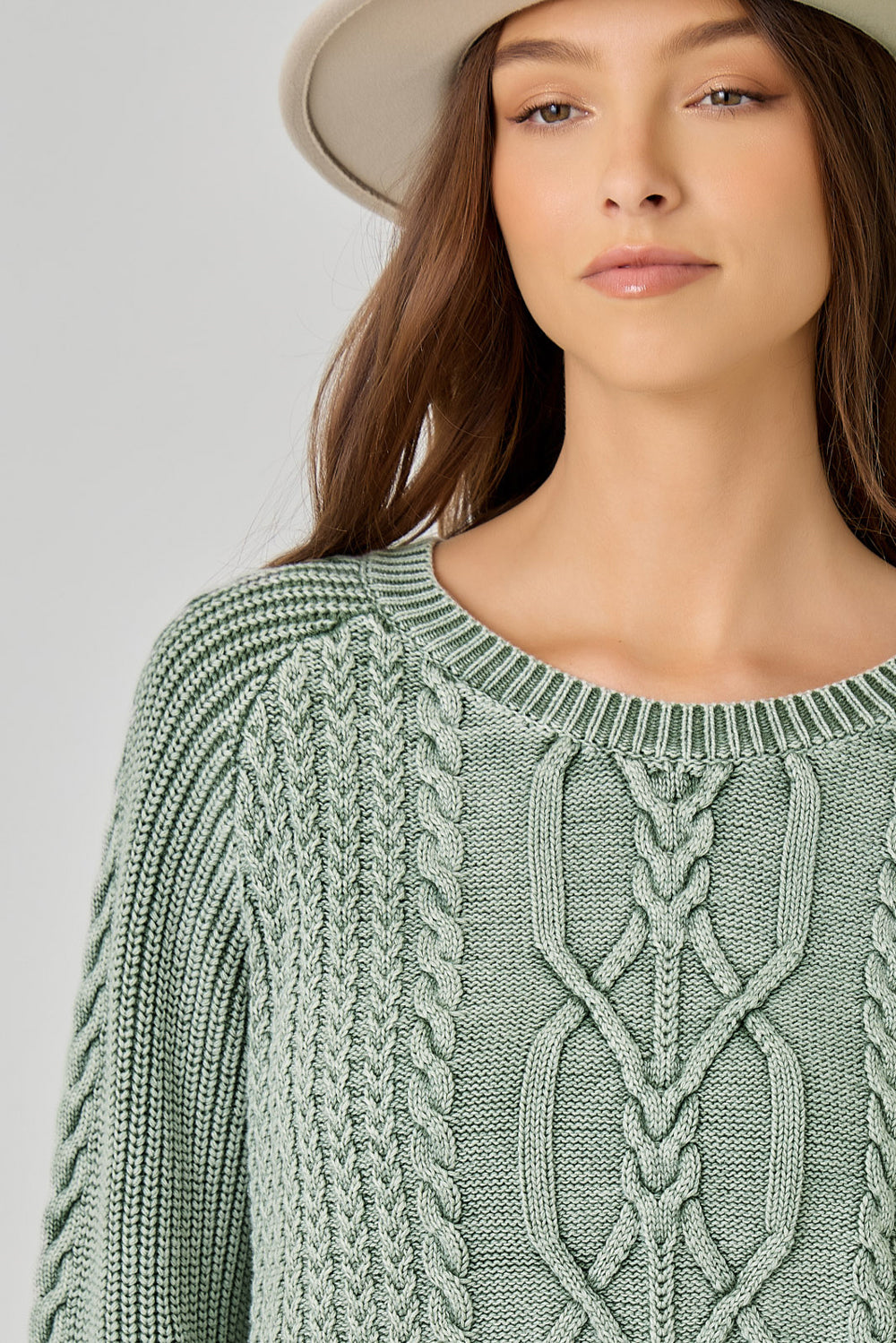 A close-up of a woman wearing a sage green cable-knit sweater and a light beige wide-brim hat against a neutral gray background.