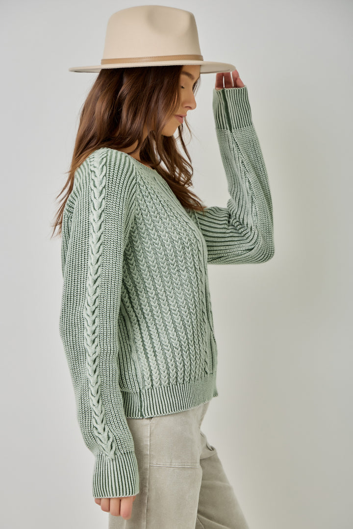 A woman wearing a sage green cable-knit sweater, light beige pants, and a beige wide-brim hat, shown from a side profile against a light gray studio background.