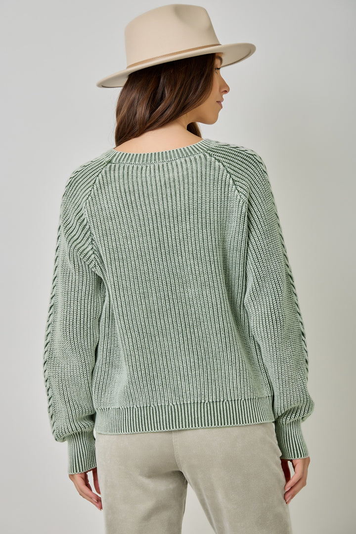 A woman shown from the back wearing a sage green ribbed knit sweater with textured sleeve details, paired with light beige pants and a beige wide-brim hat, against a neutral gray studio background.