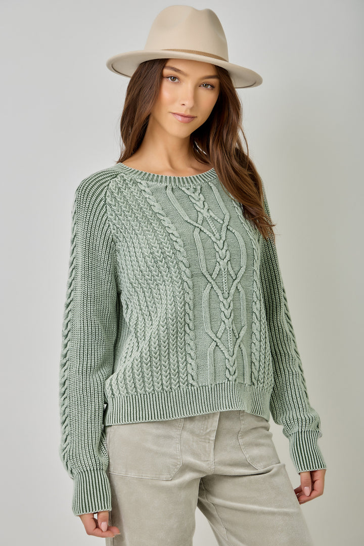 A woman wearing a sage green cable-knit sweater, light beige pants, and a beige wide-brim hat, photographed against a neutral light gray studio background.