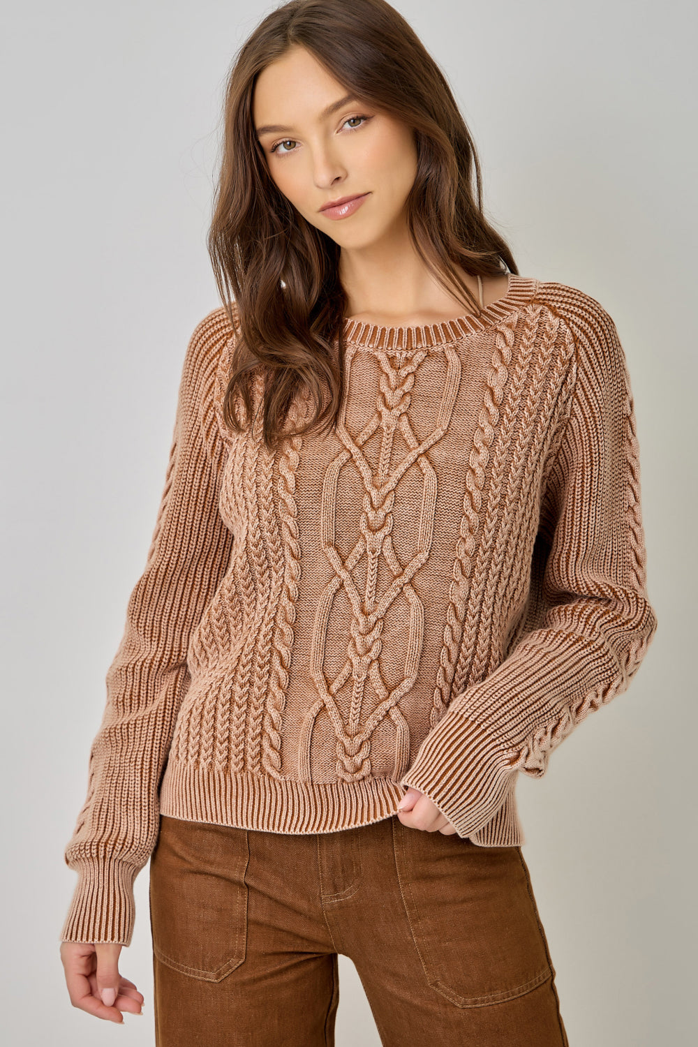A woman wearing a chunky cable-knit crewneck sweater in a warm taupe-brown color, paired with brown corduroy pants, against a neutral light gray background.