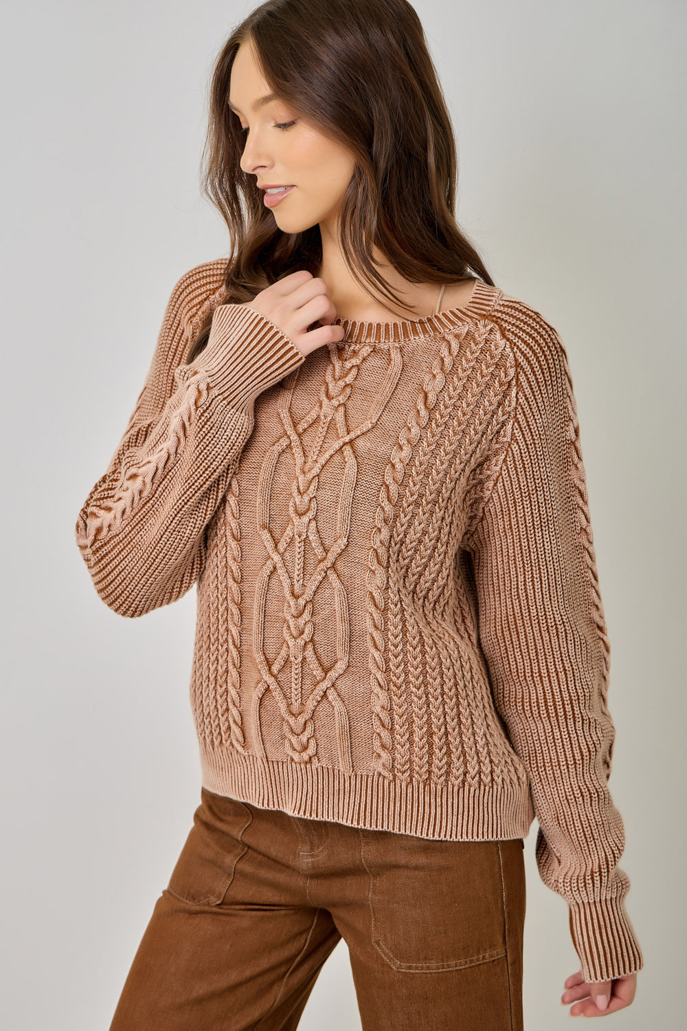 A woman wearing a chunky cable-knit sweater in a warm taupe-brown color, paired with brown pants, photographed against a neutral light gray studio background.