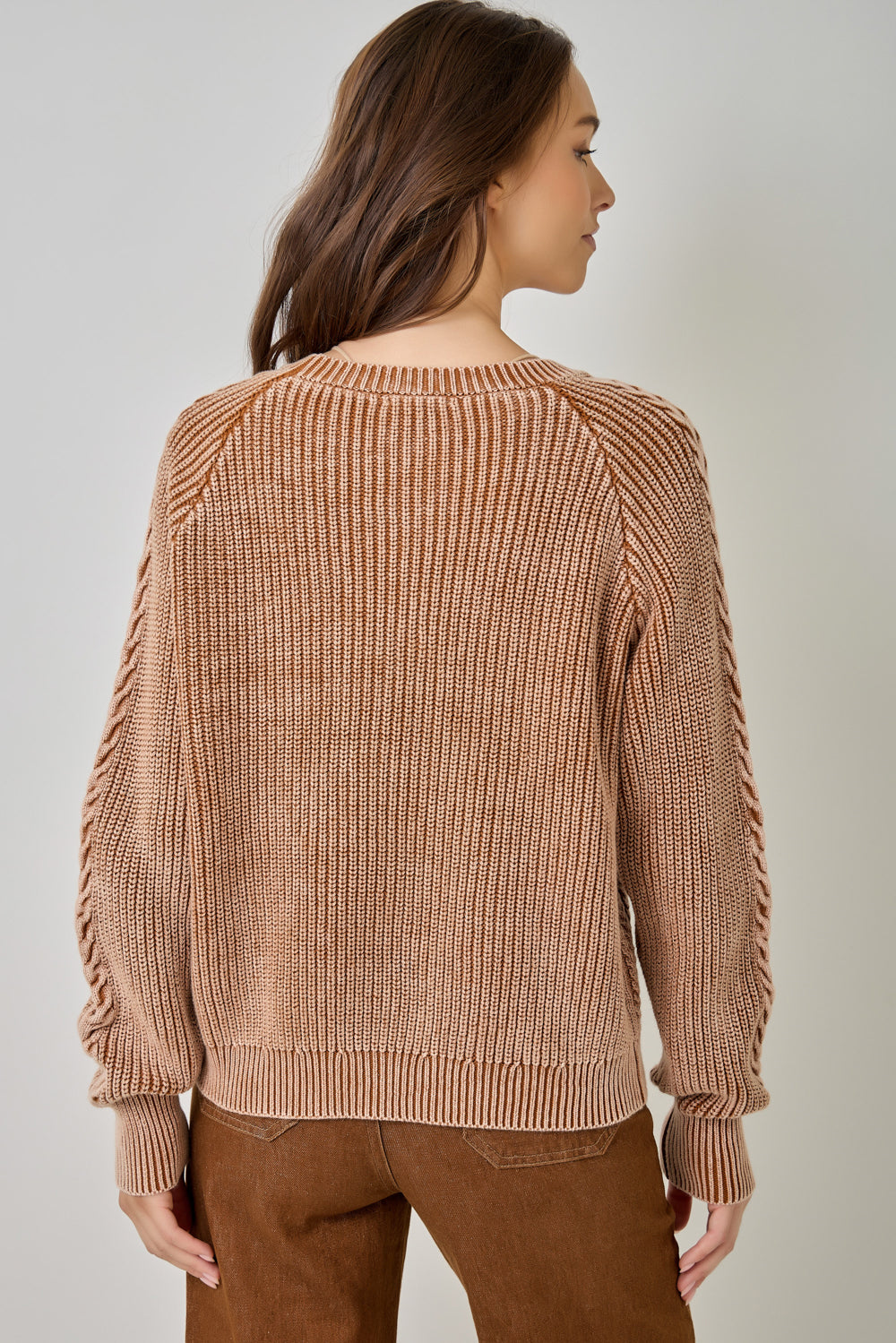A woman shown from the back wearing a ribbed knit sweater in a warm tan-brown color, paired with brown pants, against a plain light gray studio background.