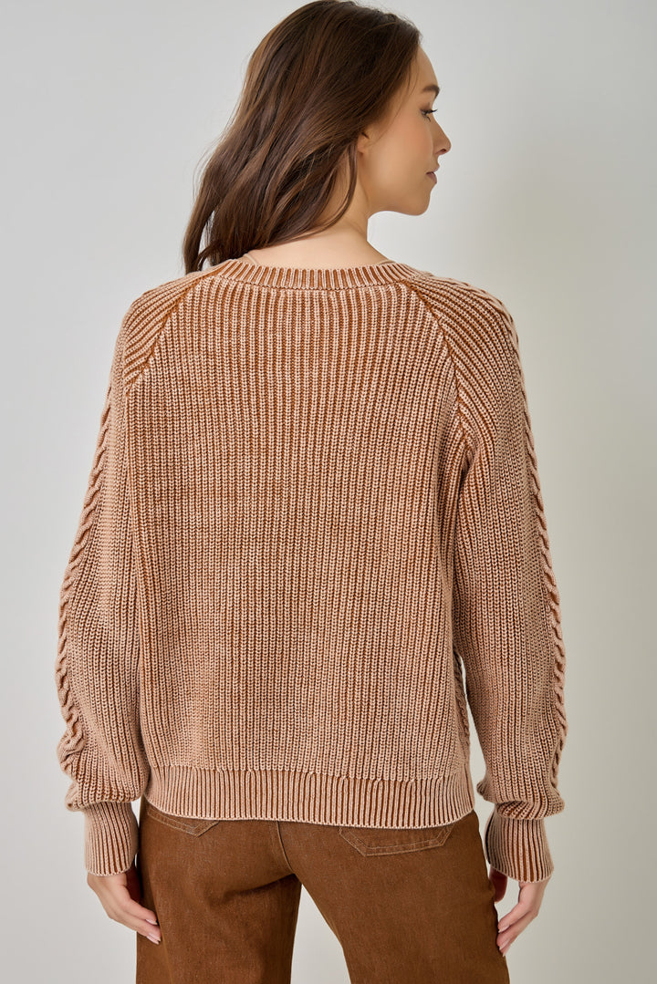 A woman shown from the back wearing a ribbed knit sweater in a warm tan-brown color, paired with brown pants, against a plain light gray studio background.