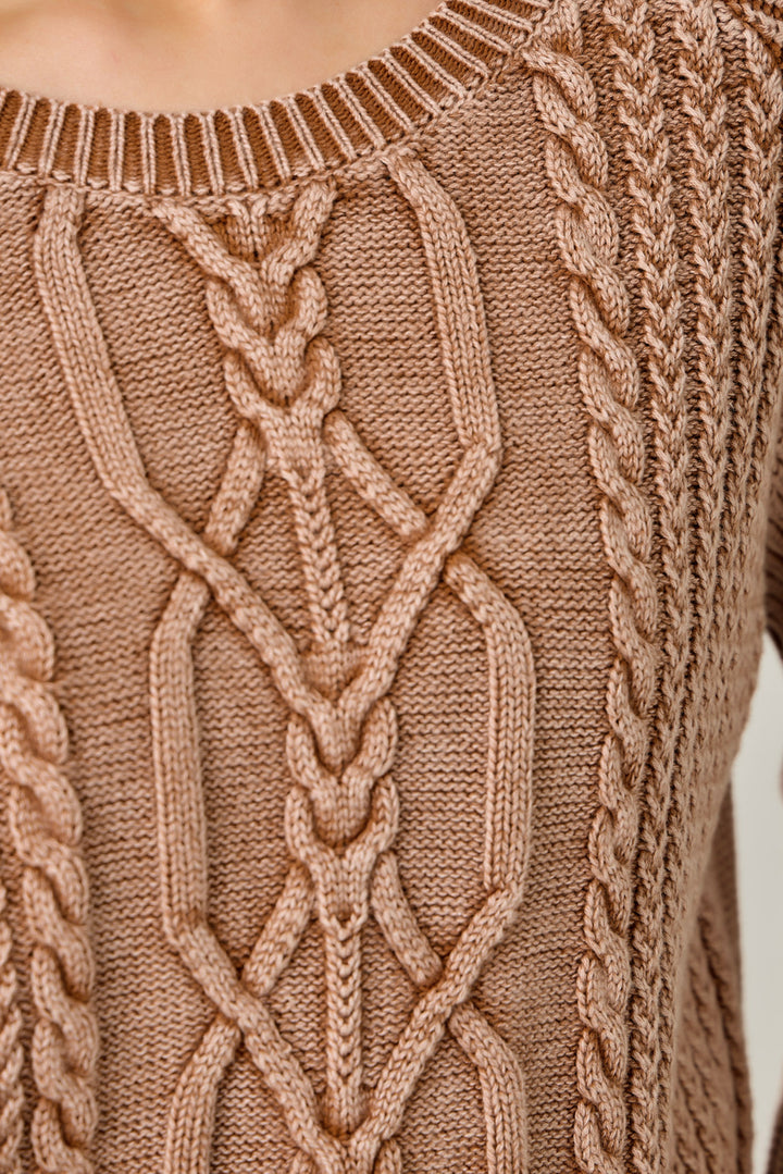 Close-up of a cable-knit sweater in a warm taupe-brown color, showing detailed knit patterns and ribbed neckline.
