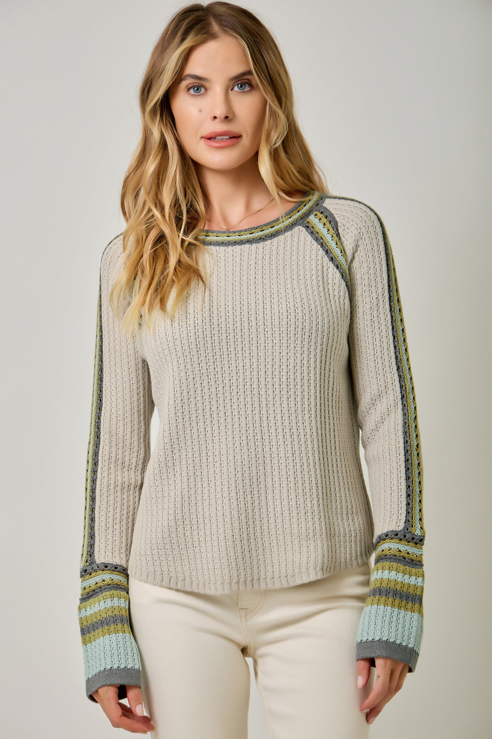 A woman wearing a cream-colored cable-knit sweater with green and yellow striped raglan sleeves and cuffs, paired with cream pants, against a light gray studio background.