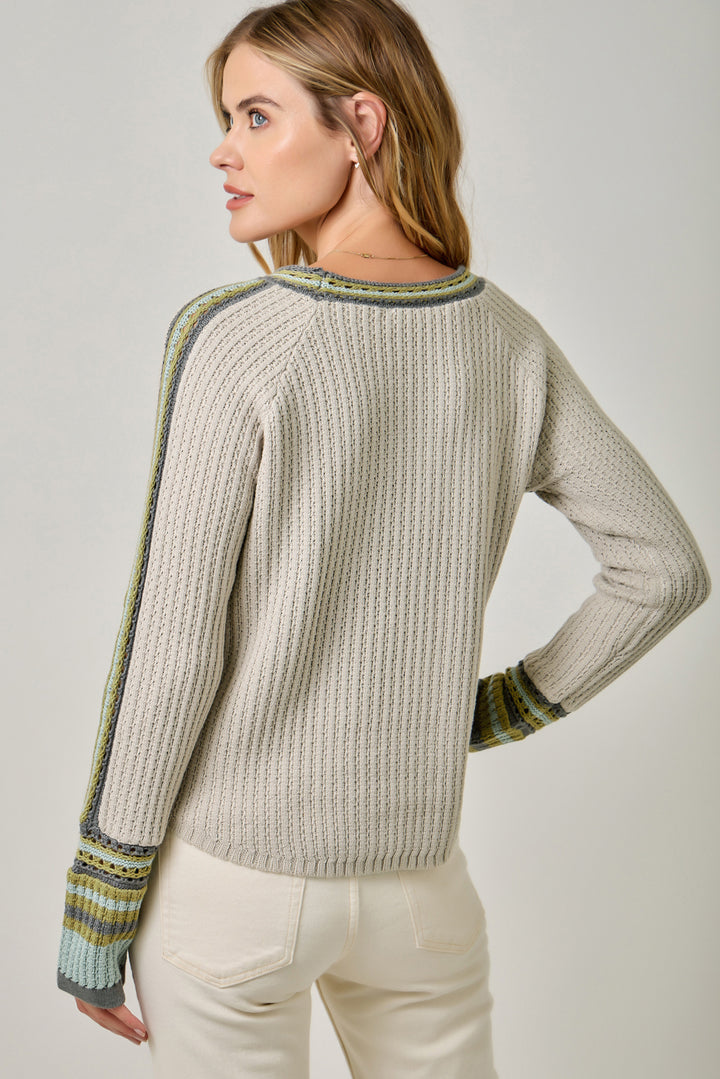 A woman wearing a light beige cable-knit sweater with green and blue striped ribbed trim on the shoulders and cuffs, shown from a three-quarter back view against a neutral studio background.