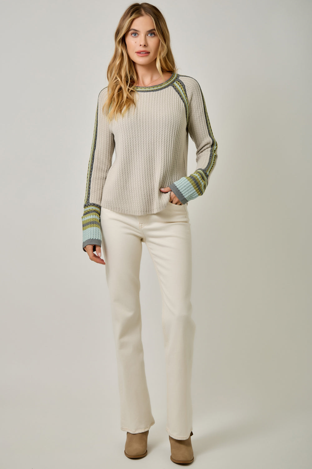 A woman modeling a ribbed knit sweater with green and blue striped raglan sleeves and cuffs, paired with cream pants, against a light gray studio background.