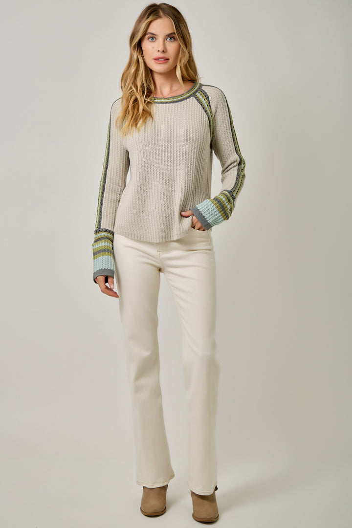 A woman modeling a ribbed knit sweater with green and blue striped raglan sleeves and cuffs, paired with cream pants, against a light gray studio background.