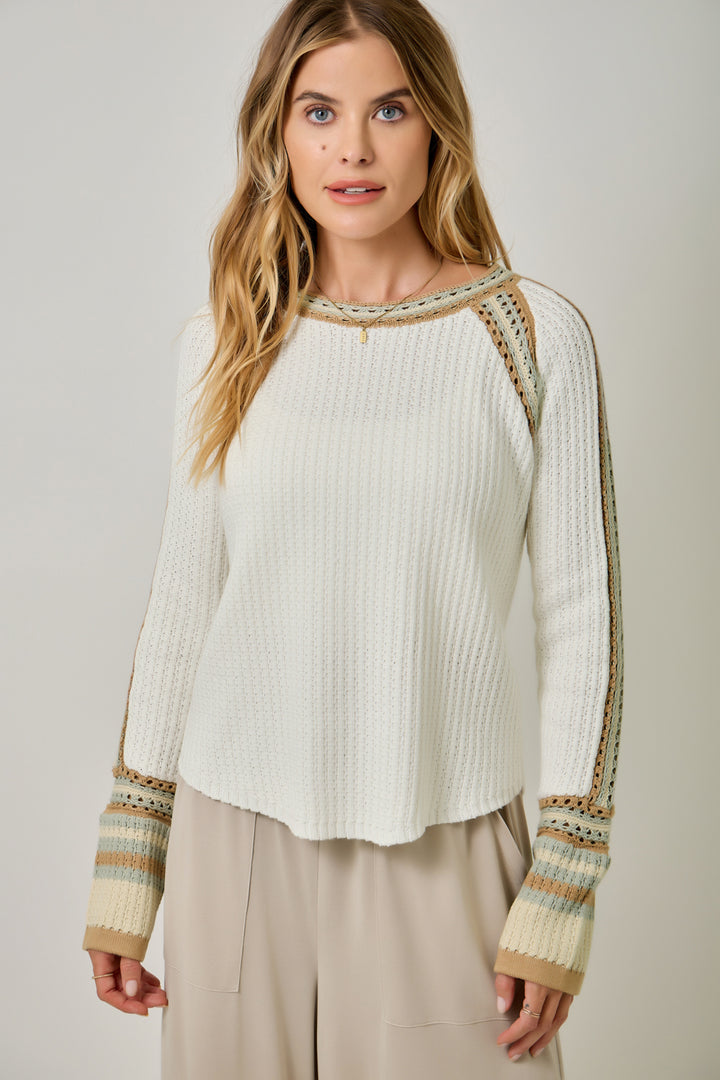 A woman wearing a cream ribbed knit long-sleeve top with patterned trim along the shoulders and cuffs, paired with beige pants, against a light gray studio background.