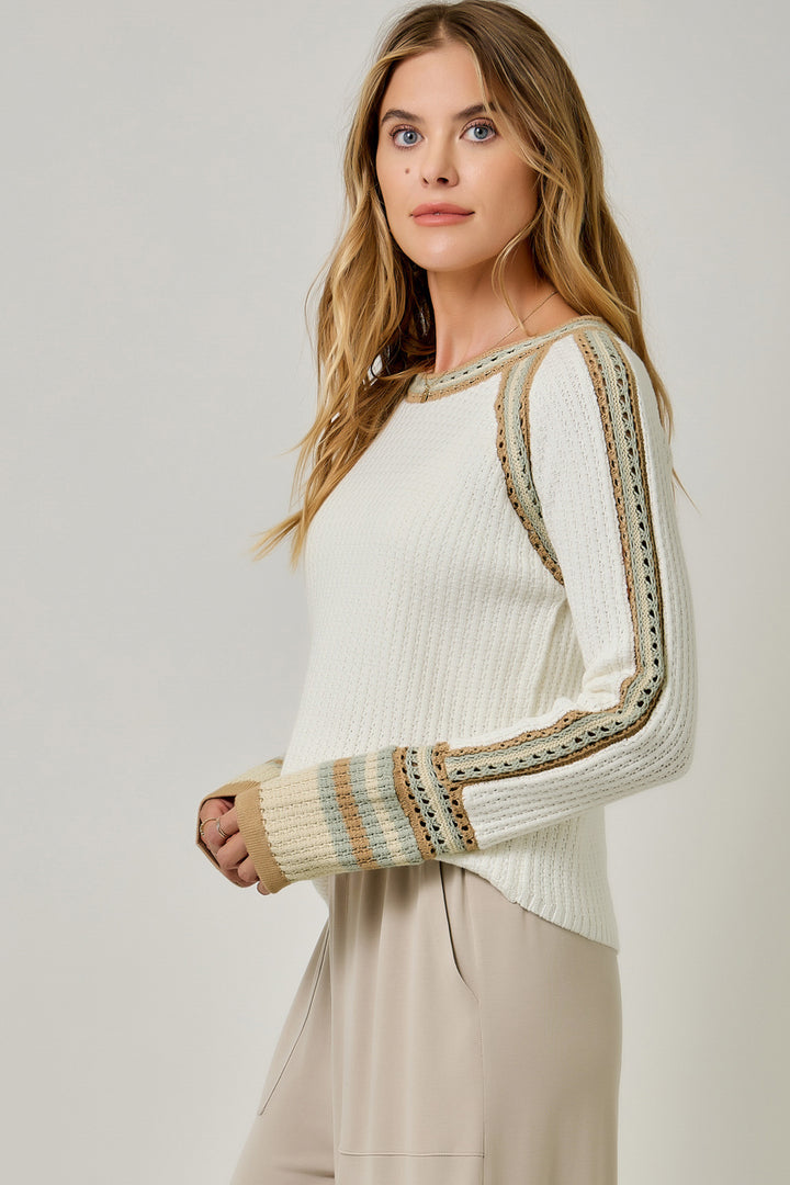 A woman modeling a cream-colored knit sweater with beige and blue striped sleeve details, paired with beige trousers, against a light gray studio background.