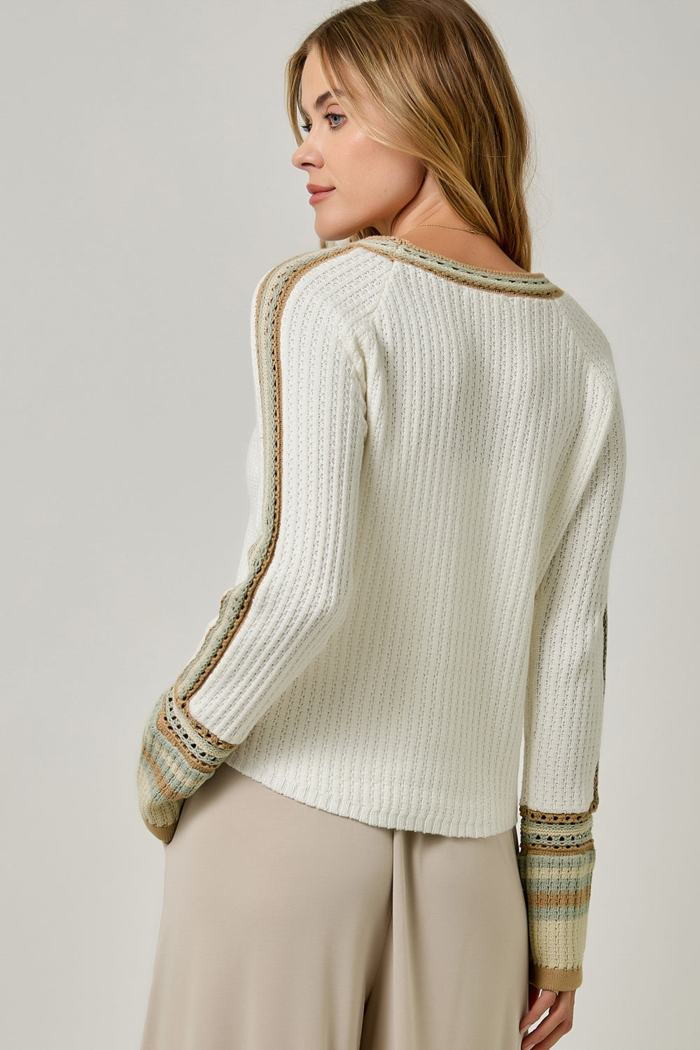 A woman shown from the back wearing a cream-colored textured knit sweater with beige and muted green patterned trim along the shoulders, cuffs, and back, paired with beige trousers against a light gray studio background.