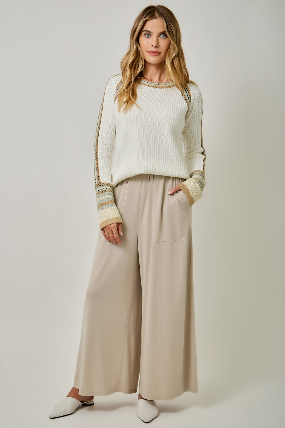 A woman modeling a cream ribbed knit sweater with gold and beige striped sleeve details, paired with wide-leg beige trousers and white pointed flats, against a light gray studio background.
