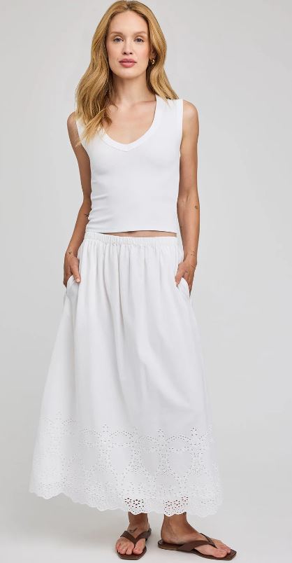 Woman wearing a white sleeveless top and long white skirt with lace details on a plain background