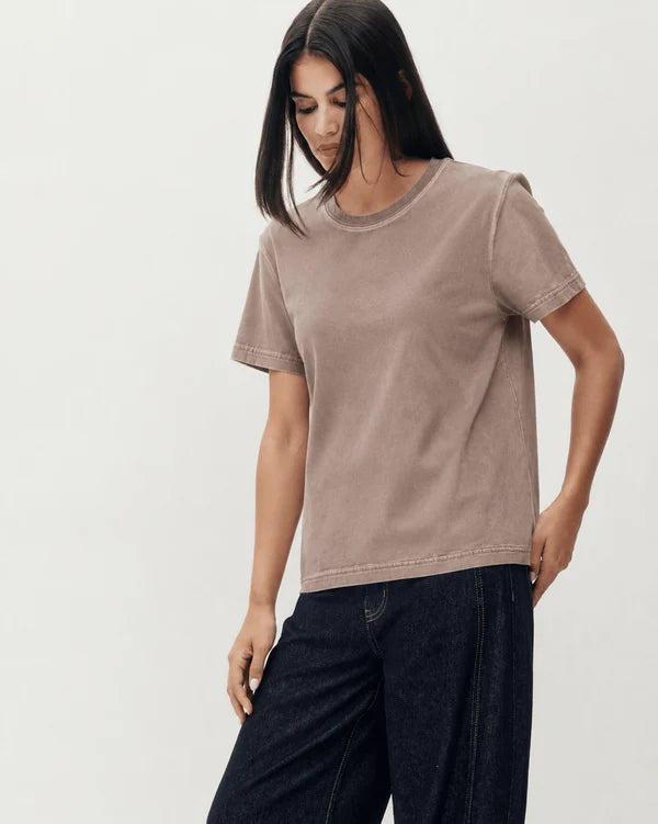 A woman wearing a short-sleeve crewneck t-shirt in a washed taupe-brown color, paired with dark wide-leg jeans, against a plain light gray studio background.