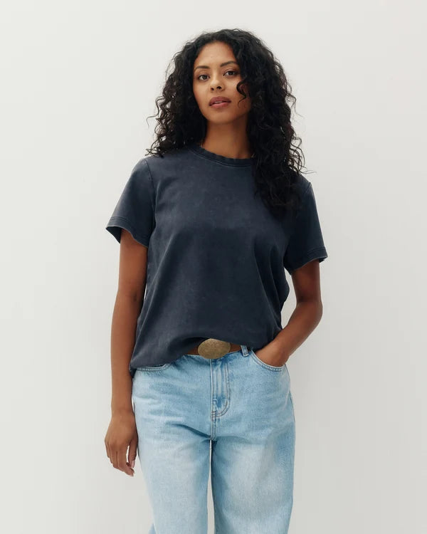 A woman with curly dark hair wearing a faded black short-sleeve crewneck t-shirt and light wash blue jeans, standing against a plain light gray background.