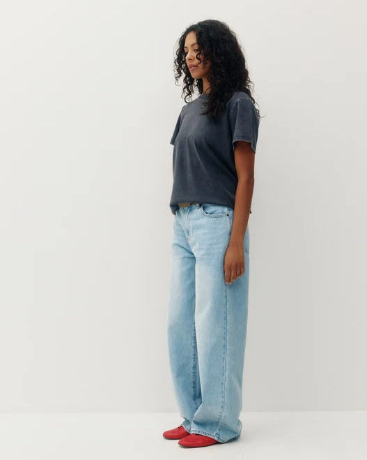 A woman stands in profile wearing a dark charcoal gray t-shirt, light-wash wide-leg blue jeans, and red slip-on shoes against a plain white studio background.