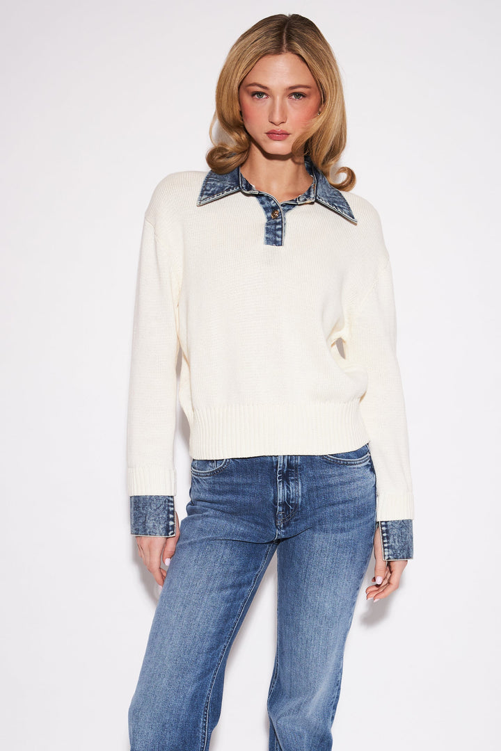 A model stands against a light gray studio background wearing a cream knit sweater with a blue denim collar and matching blue denim cuffs, paired with blue jeans and leopard-print shoes.
